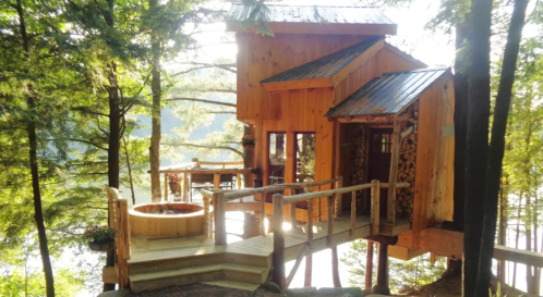 A wooden treehouse with a deck, surrounded by trees and a scenic view, featuring a fire pit and outdoor seating.
