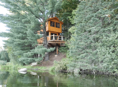 A wooden treehouse perched among tall trees, overlooking a calm lake surrounded by lush greenery.