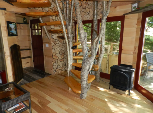 A unique spiral staircase made of tree branches in a cozy wooden interior with large windows and a small stove.