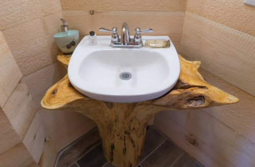 A unique wooden sink with a natural edge, set against a rustic wooden wall, featuring a soap dispenser and a bar of soap.