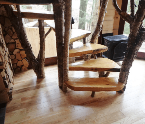 A rustic spiral staircase made of wooden slabs and tree branches, leading up in a cozy, natural setting.
