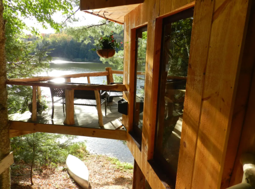 A cozy wooden cabin overlooking a serene lake, with a deck and outdoor seating surrounded by trees.