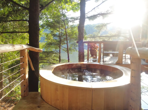 A wooden hot tub on a deck surrounded by trees, with sunlight streaming through the branches.