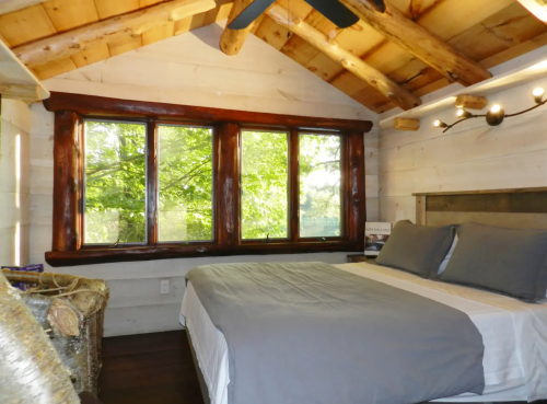 Cozy bedroom with a wooden ceiling, large windows, and a neatly made bed with gray bedding. Natural light fills the space.
