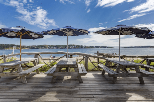 A scenic view of a beach with picnic tables and umbrellas, overlooking the water and distant shoreline under a blue sky.