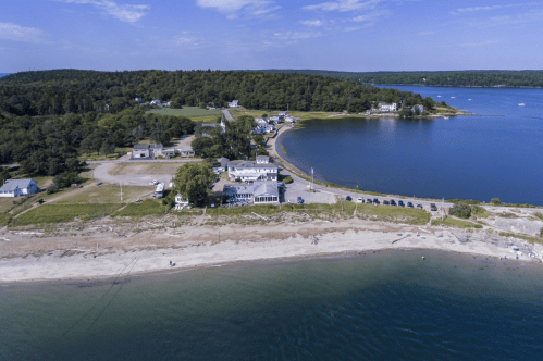 Aerial view of a coastal area with a beach, houses, and a winding road along the water under a clear blue sky.