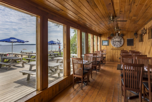 Cozy wooden interior of a restaurant with tables, large windows overlooking a beach and outdoor seating area.
