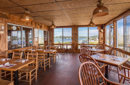 Cozy wooden restaurant interior with tables and chairs, large windows overlooking a scenic view of water and boats.