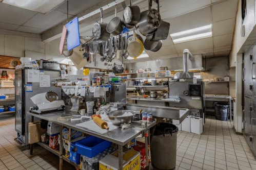 A busy kitchen with stainless steel counters, pots hanging, and various cooking equipment and ingredients scattered around.
