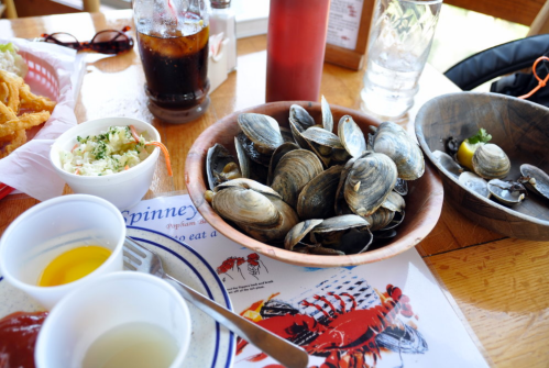 A wooden bowl filled with clams, accompanied by condiments and drinks on a table with a seafood menu.