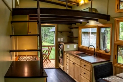 Cozy interior of a tiny house featuring a kitchen, dining area, and a staircase leading to a loft. Natural light throughout.