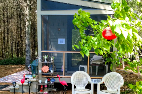 A cozy cabin surrounded by trees, featuring a screened porch, colorful flamingo decorations, and a red lantern.