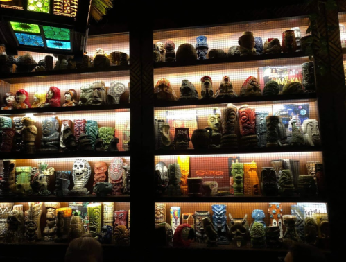 A display of colorful tiki mugs arranged on shelves, illuminated with warm lighting.