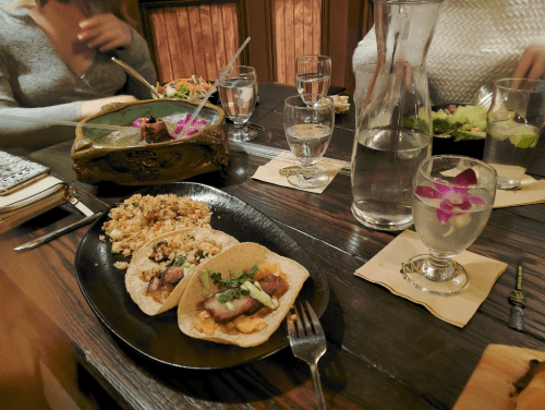 A table set with tacos, rice, salads, and drinks, featuring a decorative water pitcher and a flower garnish.