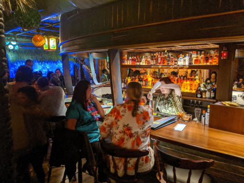 A lively bar scene with patrons enjoying drinks, colorful decor, and shelves lined with various bottles.