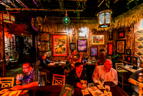 A vibrant tiki bar scene with patrons enjoying drinks, surrounded by tropical decor and colorful artwork.