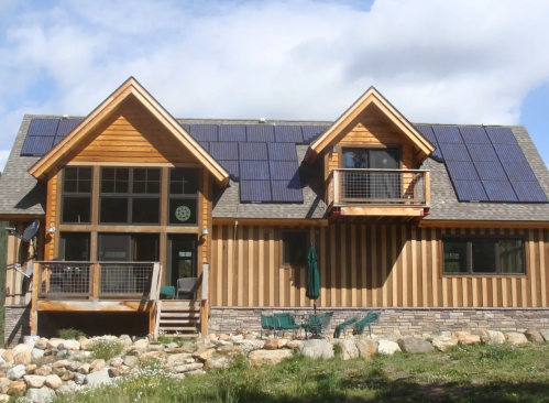 A modern wooden house with solar panels on the roof, featuring a balcony and stone landscaping in front.