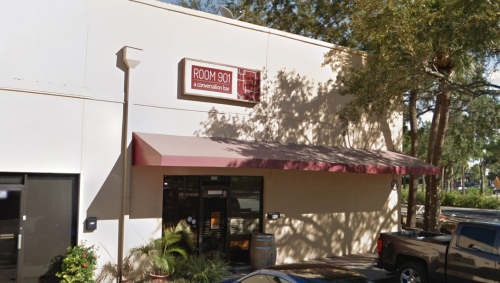 Exterior of Room 901, a conversation bar, featuring a maroon awning and a sign above the entrance. Trees in the background.