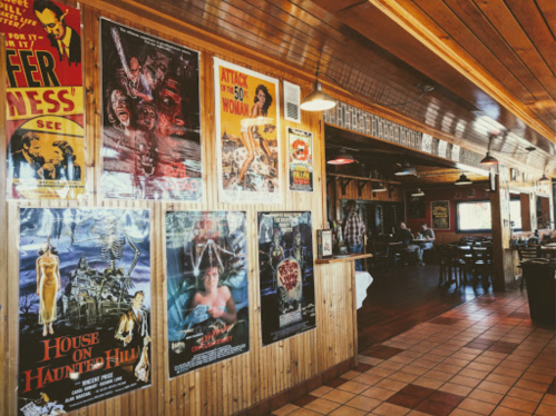 A restaurant interior featuring vintage horror movie posters on wooden walls and a cozy dining area in the background.