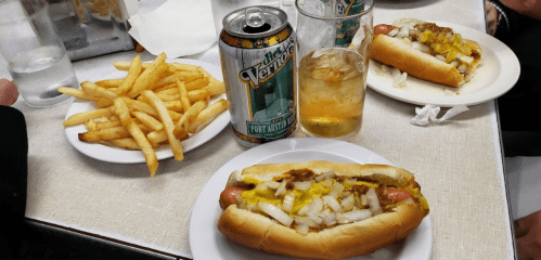 A table with two hot dogs topped with mustard and onions, a plate of fries, a can of soda, and glasses of drink.
