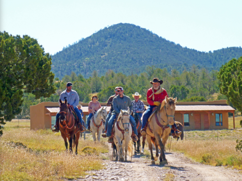 A group of six people on horseback riding down a dirt path with mountains and a building in the background.