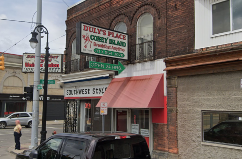 A street view of Duly's Place, a 24-hour diner, with a sign and nearby shops in an urban setting.