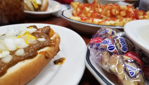 A plate with a chili dog topped with onions, alongside fries with ketchup and a pack of oyster crackers.