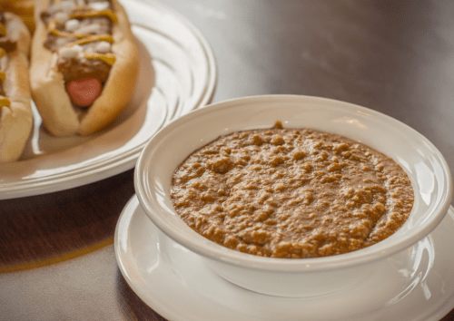 A bowl of chili sits next to a plate of hot dogs topped with mustard and onions.