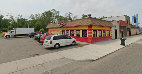 A colorful fast-food restaurant with a red and yellow exterior, parked cars, and a delivery truck nearby.