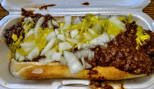 A hot dog in a foam container topped with chili, diced onions, and mustard.