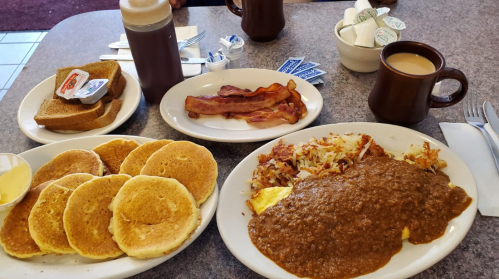 A breakfast spread featuring pancakes, bacon, hash browns, a meat sauce omelet, toast, and coffee.