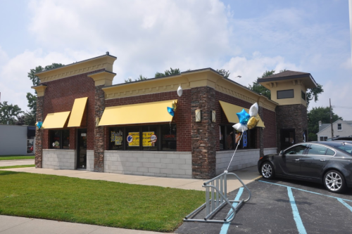 A brick restaurant with yellow awnings and decorative balloons outside, featuring a parking area and grassy lawn.