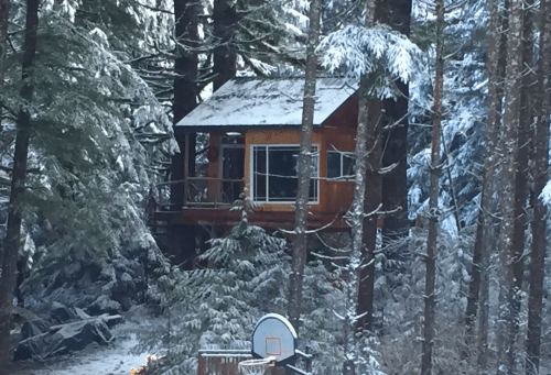 A cozy wooden cabin nestled among snow-covered trees, with a basketball hoop in the foreground.