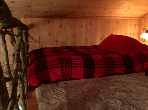Cozy loft bedroom with a red and black plaid blanket, wooden walls, and a soft rug. A lamp provides warm lighting.