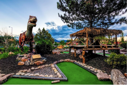 A mini-golf course featuring a large dinosaur statue, a thatched-roof hut, and lush greenery under a blue sky.