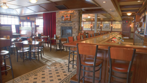 Cozy restaurant interior with wooden bar, tables, and a stone fireplace, featuring warm lighting and a welcoming atmosphere.