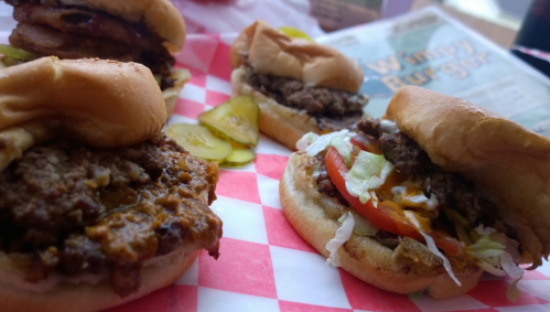 A close-up of two burgers on a red and white checkered paper, one with pickles and the other with lettuce and tomato.