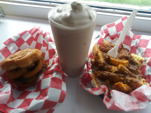 A burger with onion rings, a plate of chili cheese fries, and a thick milkshake in a glass with a straw.