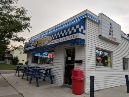 A small diner with a blue and white checkered awning, featuring signs for hot dogs, shakes, and open hours.