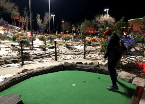 A person in a red beanie prepares to putt on a mini-golf course decorated with colorful lights at night.