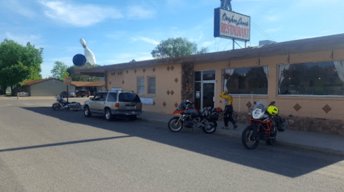 A restaurant with a retro sign, parked motorcycles, and a person in a yellow shirt outside on a sunny day.