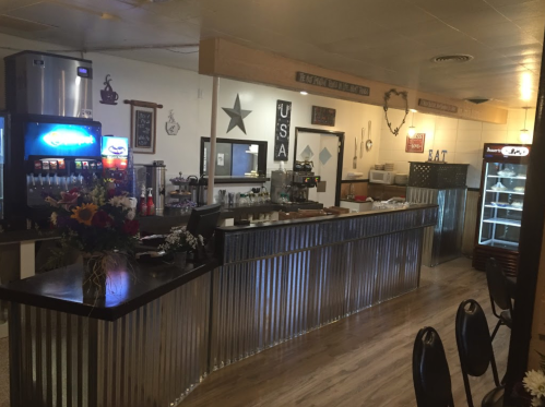 A cozy diner interior featuring a metal counter, drink dispensers, and a display fridge, with rustic decor and seating.