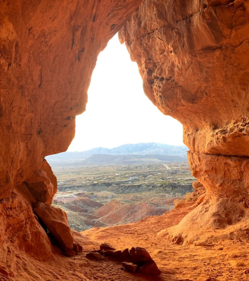 View from a cave opening, showcasing a landscape of red rock formations and distant mountains under a bright sky.