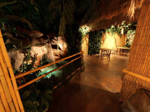 A tropical-themed indoor space with a waterfall, lush greenery, and bamboo furniture.