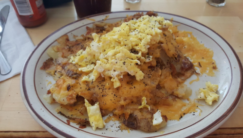 A plate of cheesy hash browns topped with scrambled eggs and black pepper, served with a drink in the background.