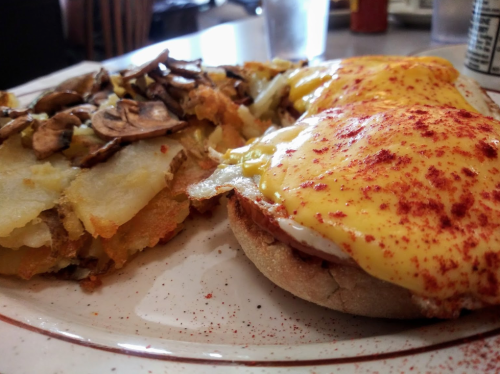 A plate of eggs Benedict with hollandaise sauce and a side of crispy hash browns topped with mushrooms.