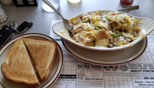 A plate of scrambled eggs with vegetables and ham, served with a side of toasted bread on a diner table.