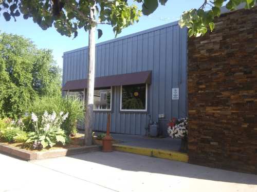 A gray building with a brown awning, surrounded by greenery and colorful flowers, on a sunny day.