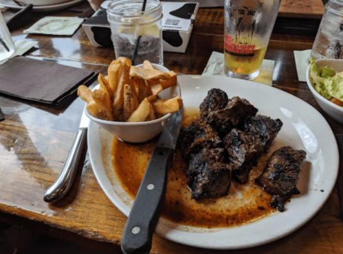 A plate of grilled meat with a side of fries, served on a wooden table with drinks in the background.