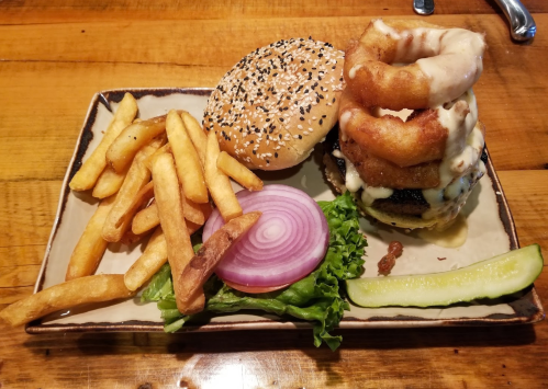 A plate with a sesame seed burger, onion rings, fries, lettuce, tomato, and a pickle on a wooden table.